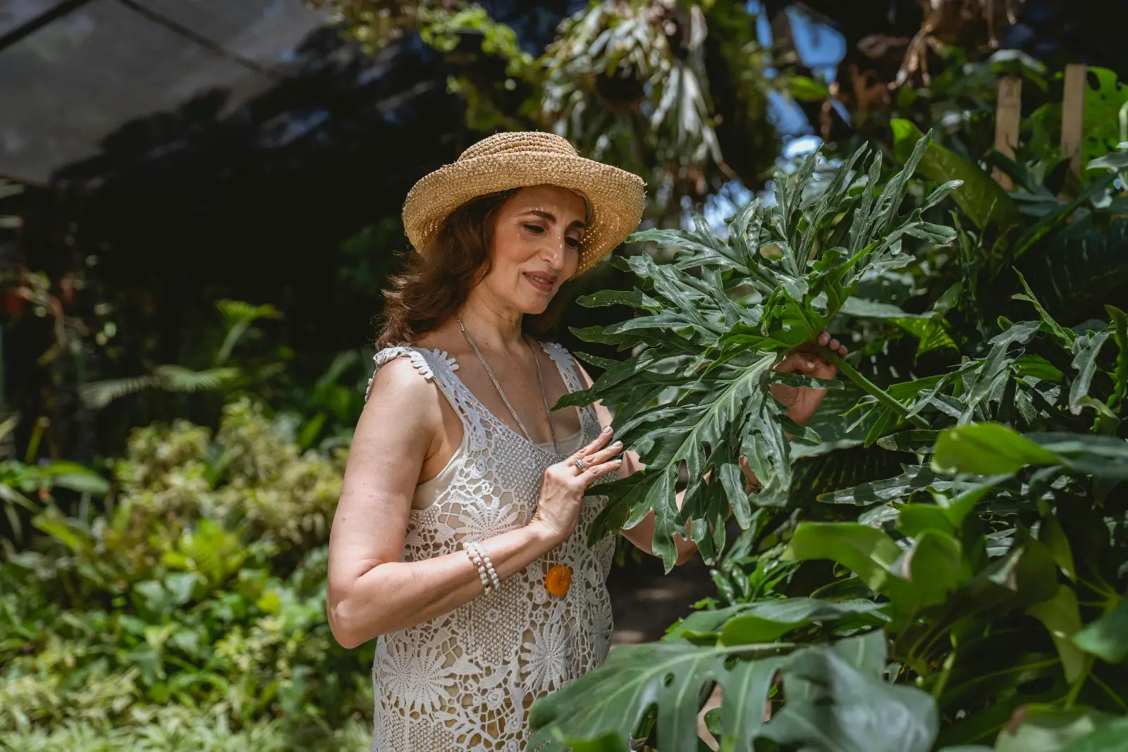 Femme avec un chapeau, une robe, les mains dans les plantes d'un jardin.