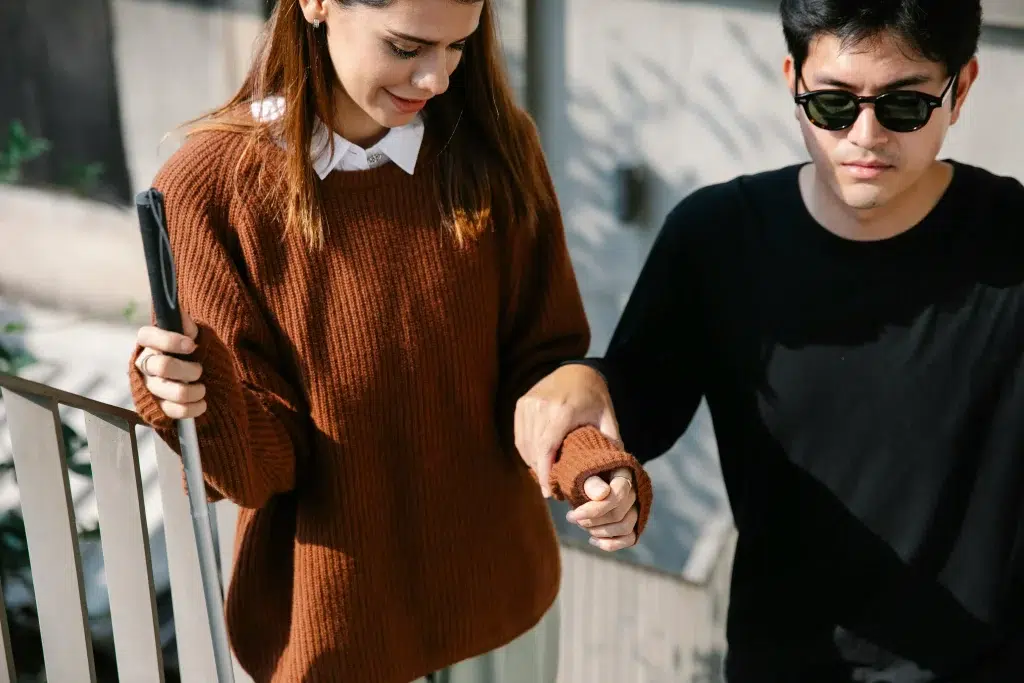 Deux personne se tenant la main. L'une porte des lunettes de soleils et atteint d'un handicap visuel.